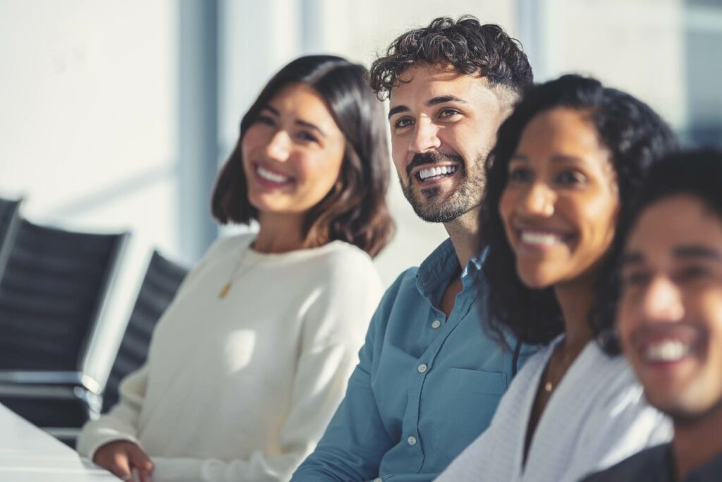 Group of people listening to a presentation. The are at a seminar or meeting sitting at a table. They are all looking to the front of the room. Men and women dressed in business attire. Multi-cultural group with African, Latino and Caucasian